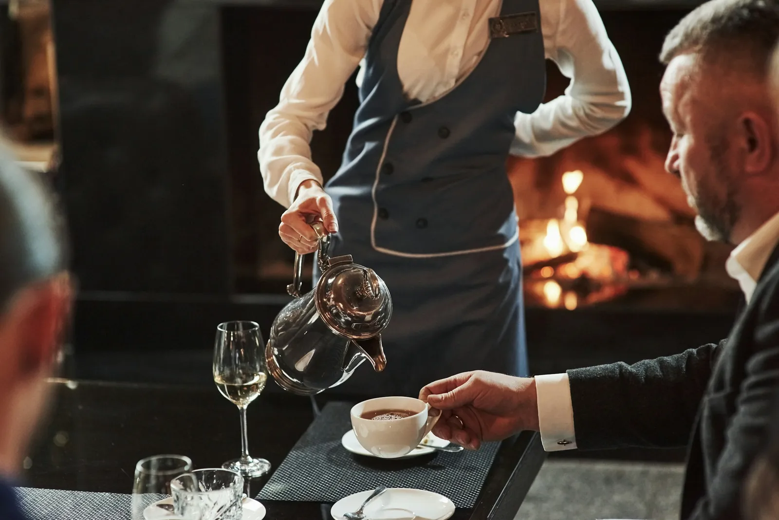 En salle, une serveuse sert un client attablé près d’une cheminée.
